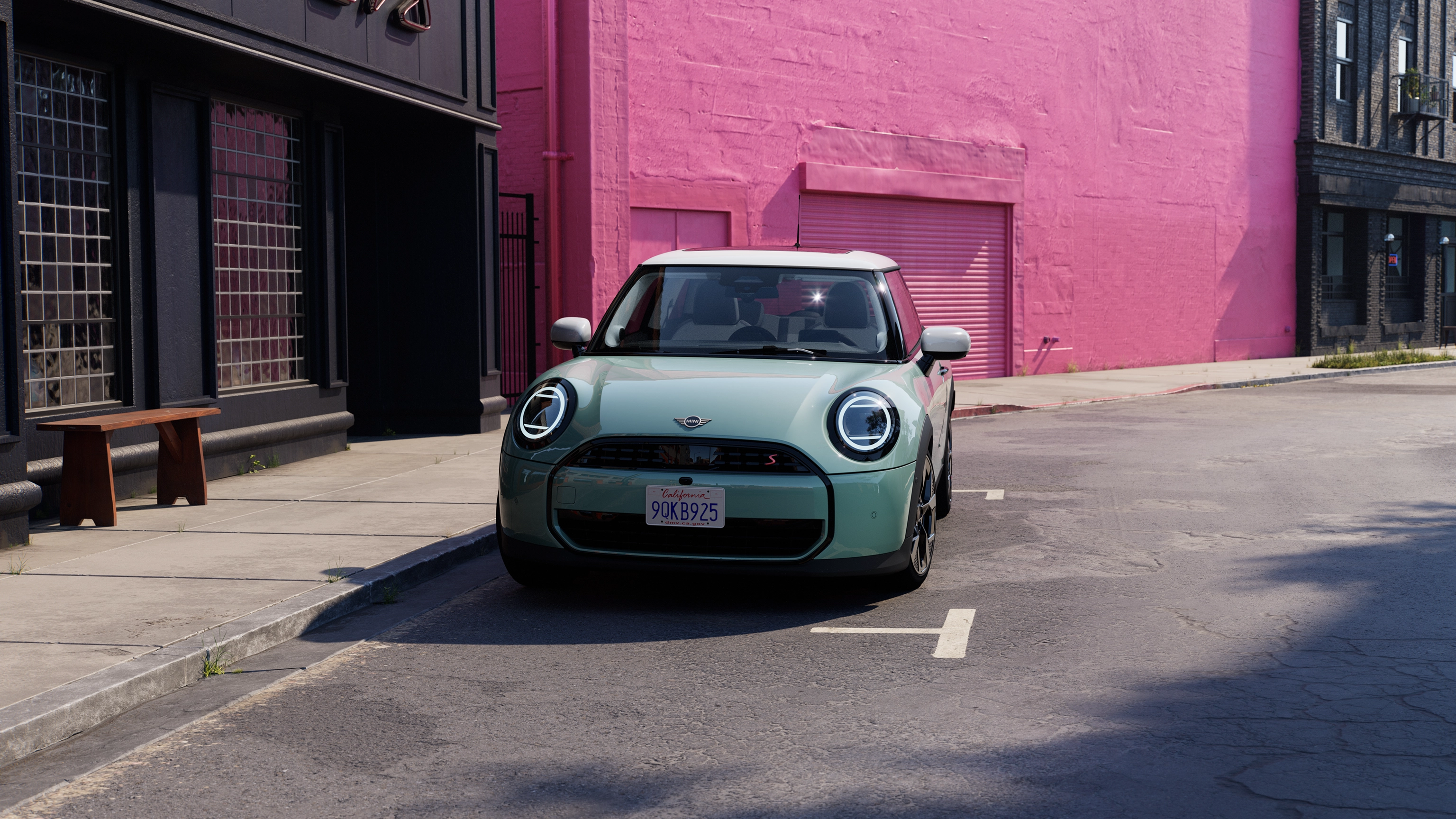 Vista frontale di una MINI Cooper in fase di parcheggio, nella colorazione Ocean Wave Green e tetto bianco su strada, con un muro rosa sullo sfondo