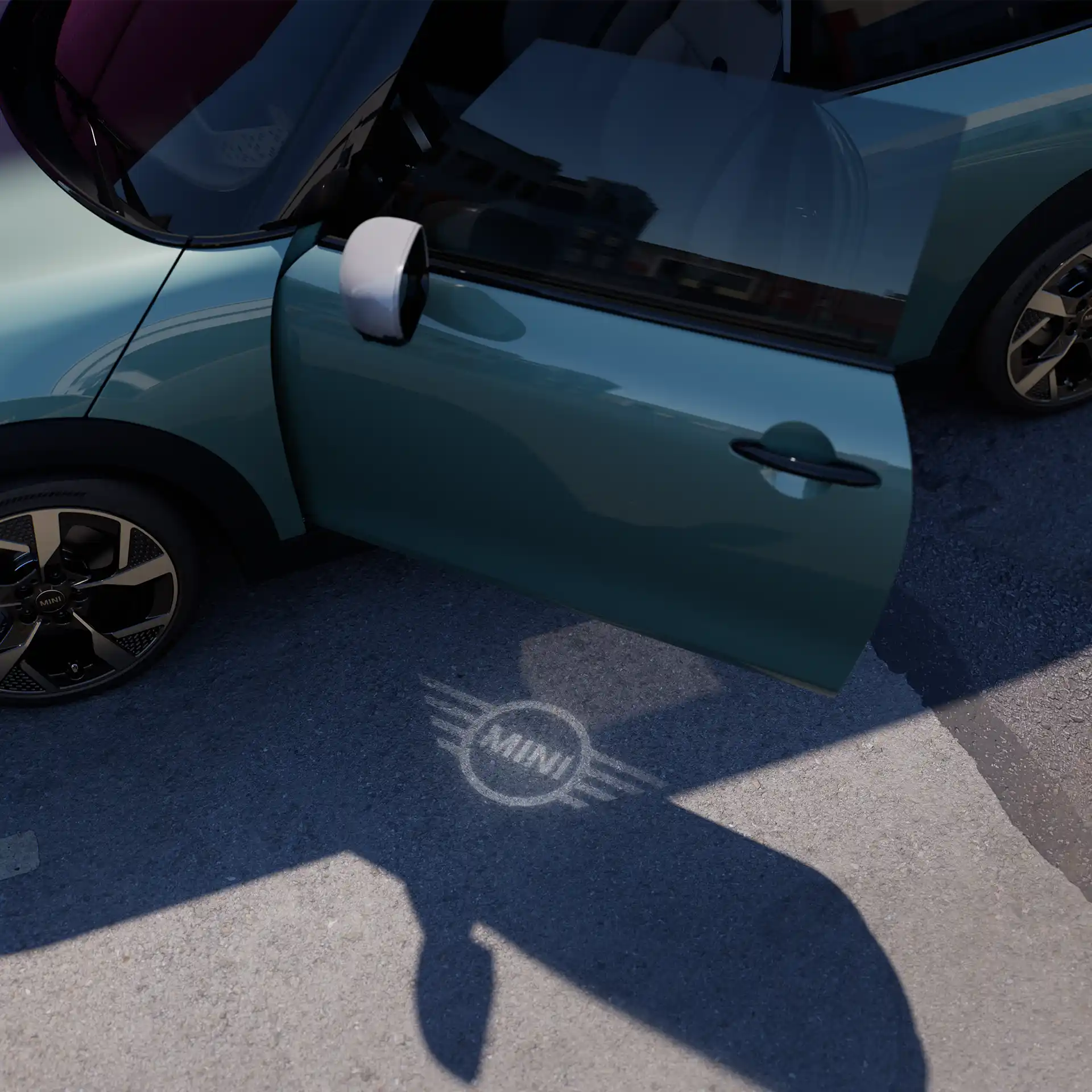 Vista esterna della MINI Cooper con portiera aperta e luce di benvenuto con il logo MINI proiettato sul suolo 