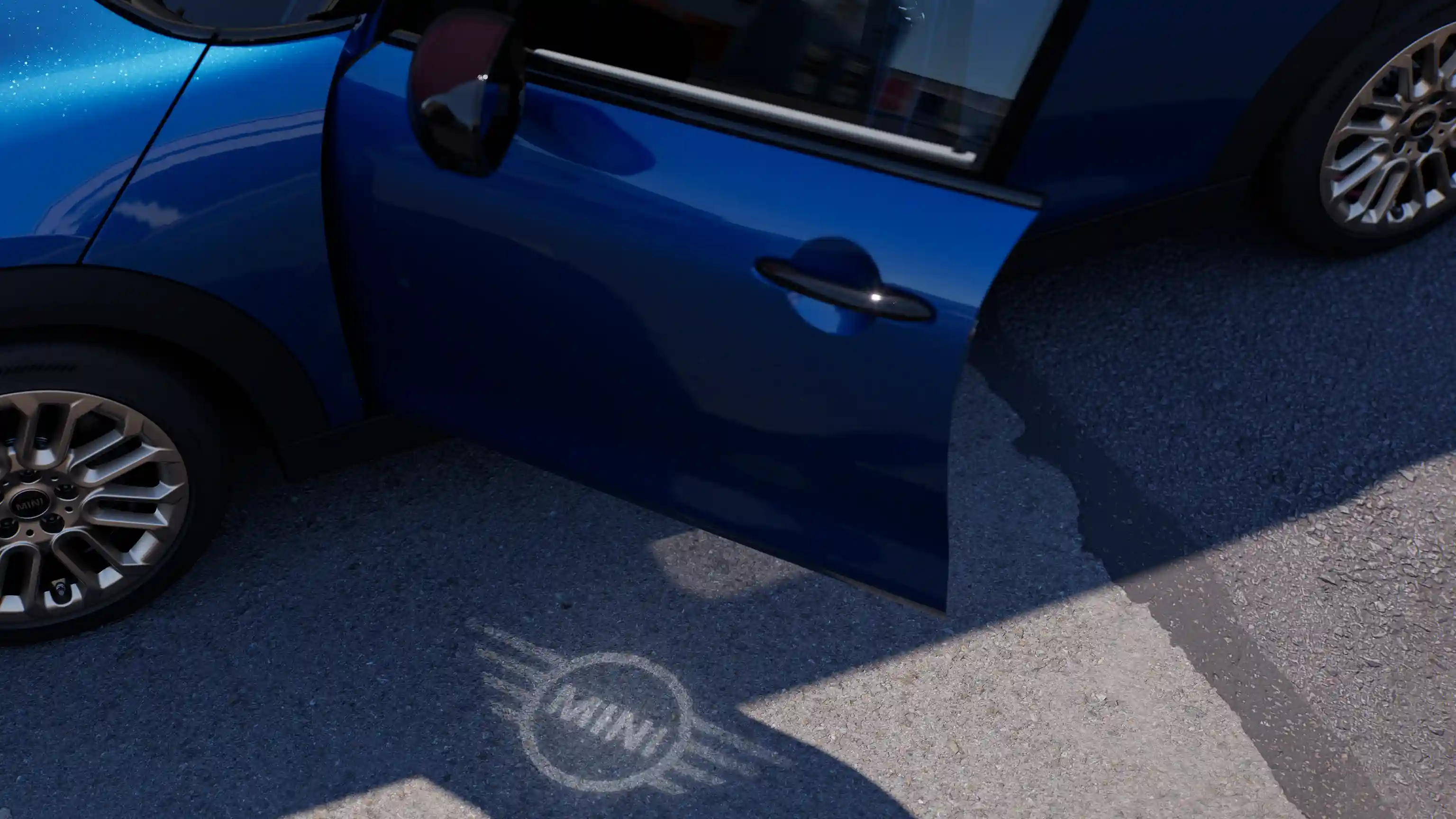 Vista esterna della MINI Cooper 5 Porte con portiera aperta e luce di benvenuto con il logo MINI proiettato sul terreno 