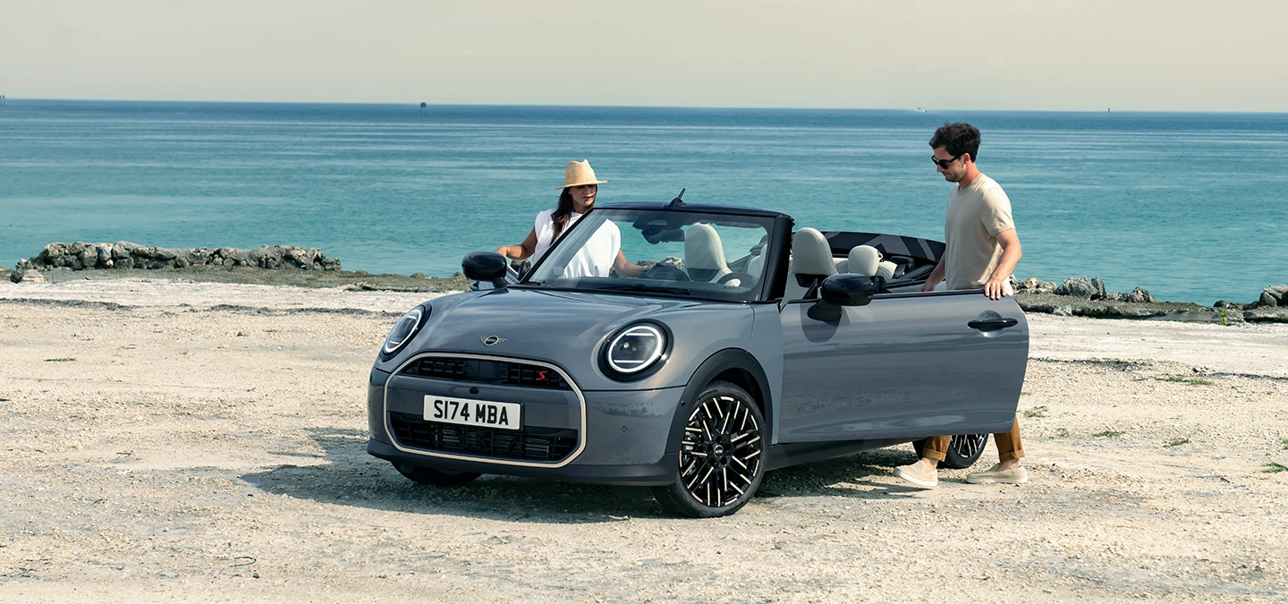 Vista frontale sinistra della MINI Cooper cabrio in Copper Grey su una spiaggia con due persone accanto.