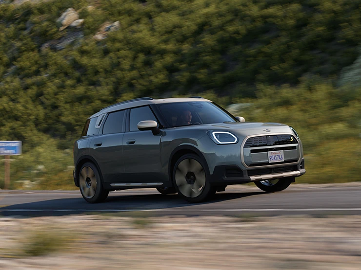 Vista laterale di MINI Countryman 100% elettrica in Smokey Green con tetto Vibrant Silver su un percorso fuoristrada.