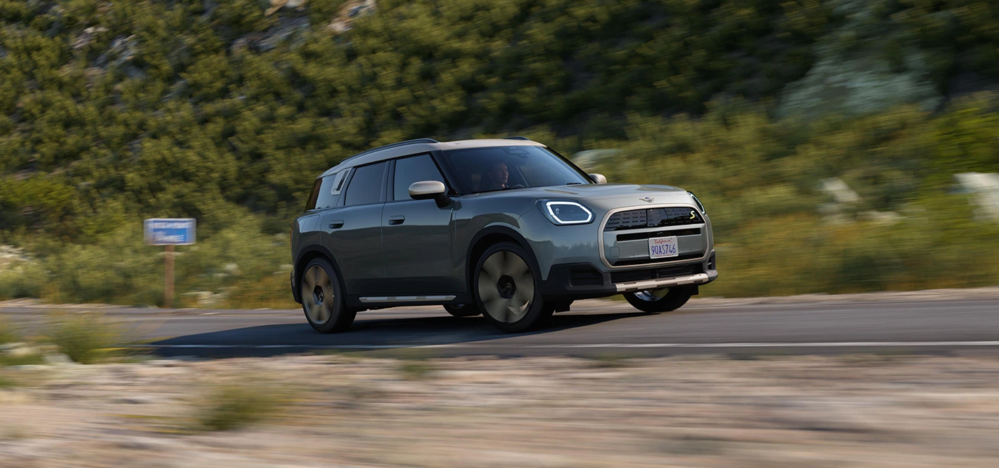 Vista laterale di MINI Countryman 100% elettrica in Smokey Green con tetto Vibrant Silver su un percorso fuoristrada.