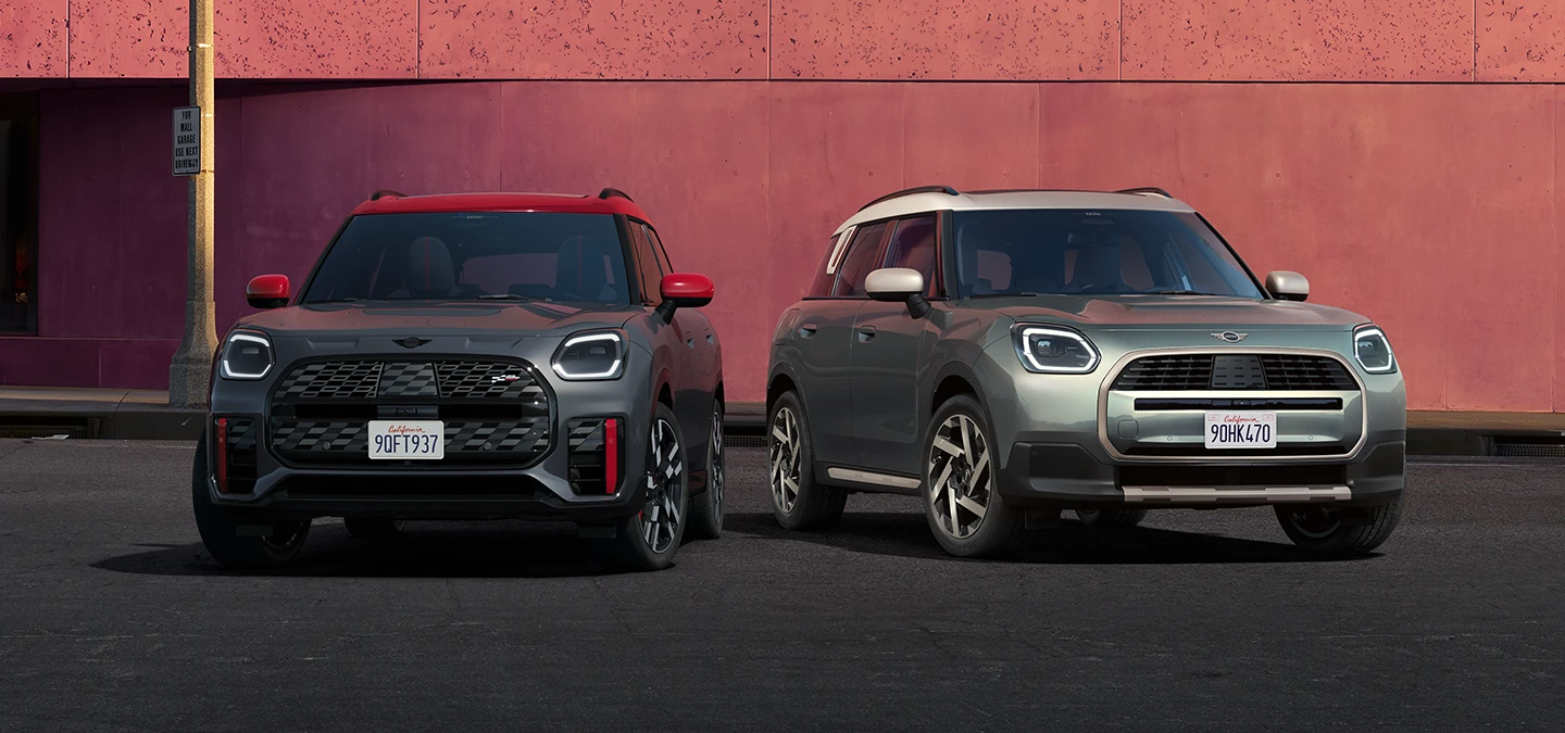 Vista frontale della MINI Countryman JCW in Legend Grey e con tetto rosso e della MINI Countryman in Smokey Green e con tetto Vibrant Silver, parcheggiate una accanto all'altra.