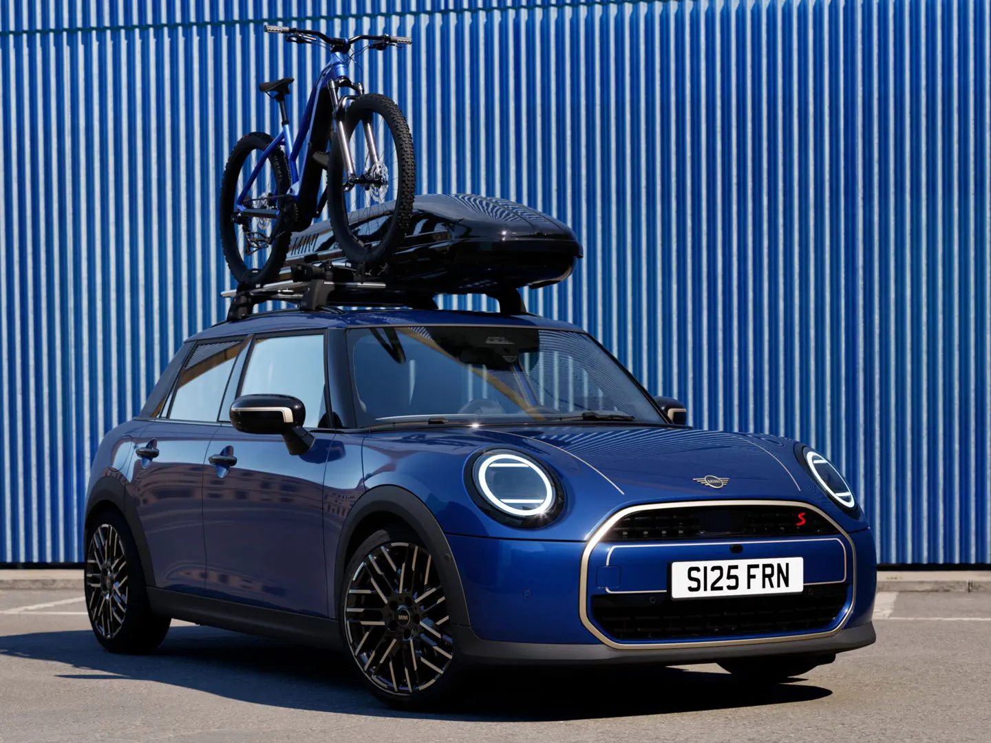 Vista frontale della MINI Aceman in Indigo Sunset Blue con portabici da tetto MINI, che trasporta una mountain bike e box per tetto MINI 320.