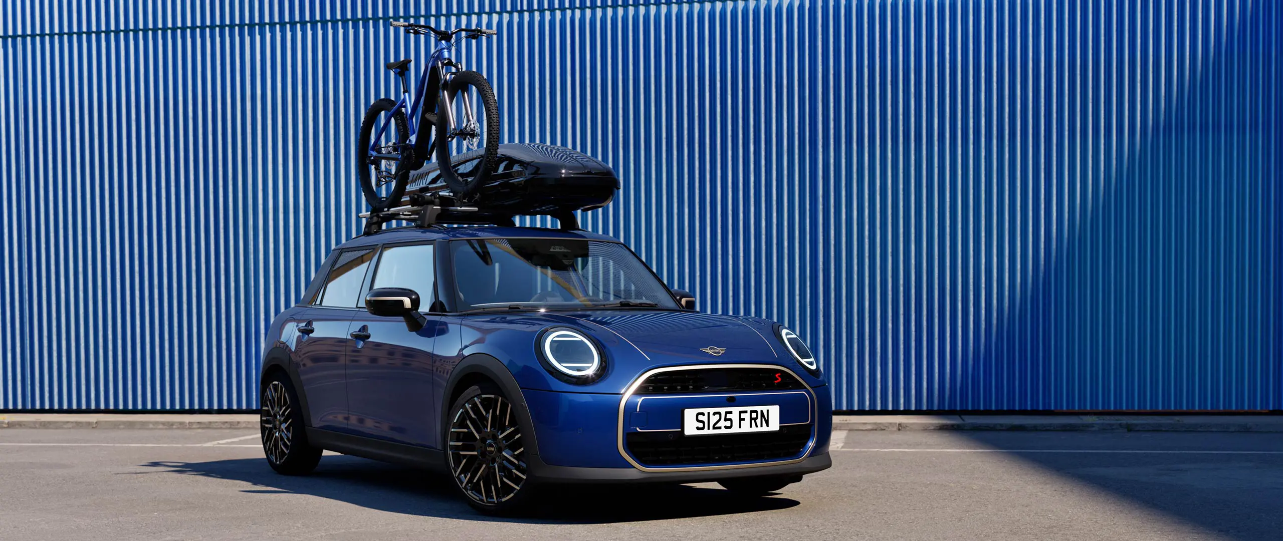 Vista frontale della MINI Aceman in Indigo Sunset Blue con portabici da tetto MINI, che trasporta una mountain bike e box per tetto MINI 320.