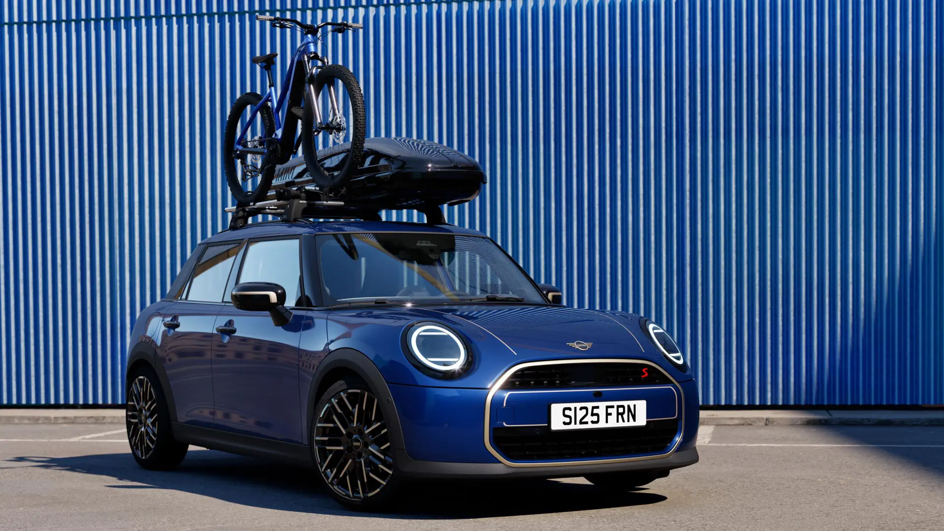 Vista frontale della MINI Aceman in Indigo Sunset Blue con portabici da tetto MINI, che trasporta una mountain bike e box per tetto MINI 320.