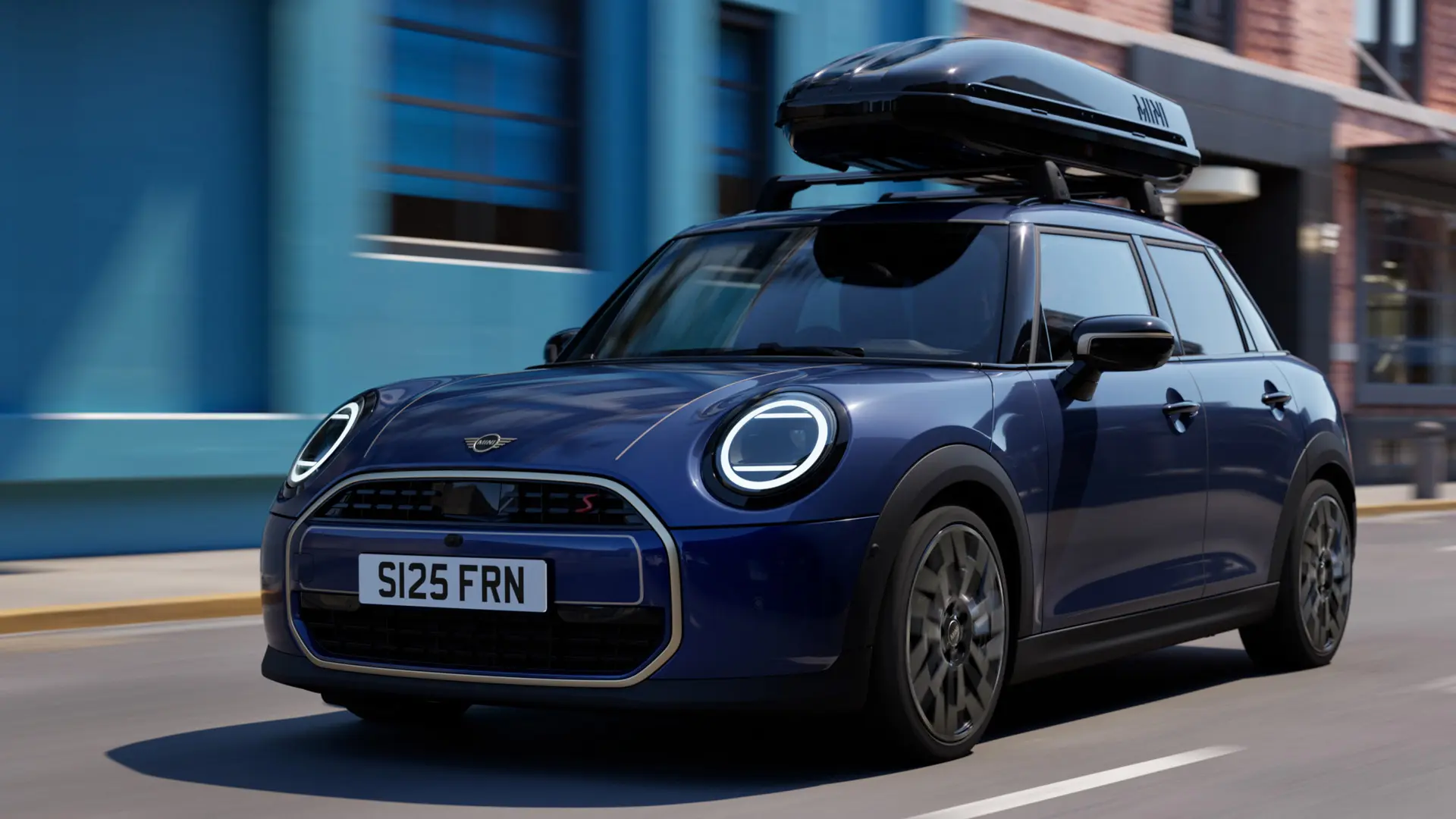 Vista frontale a tre quarti della MINI Aceman in Indigo Sunset Blue dotata di portapacchi MINI, che trasporta un box per tetto MINI 320. 
