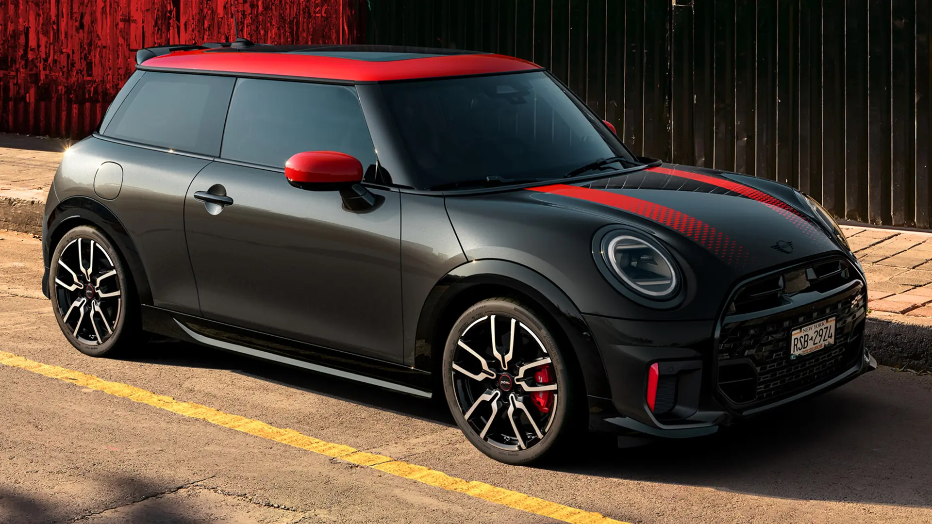 MINI John Cooper Works a 3 porte in Midnight Black con tetto e gusci retrovisori in Chili Red e strisce sportive rosse, vista laterale, parcheggiata in strada.
