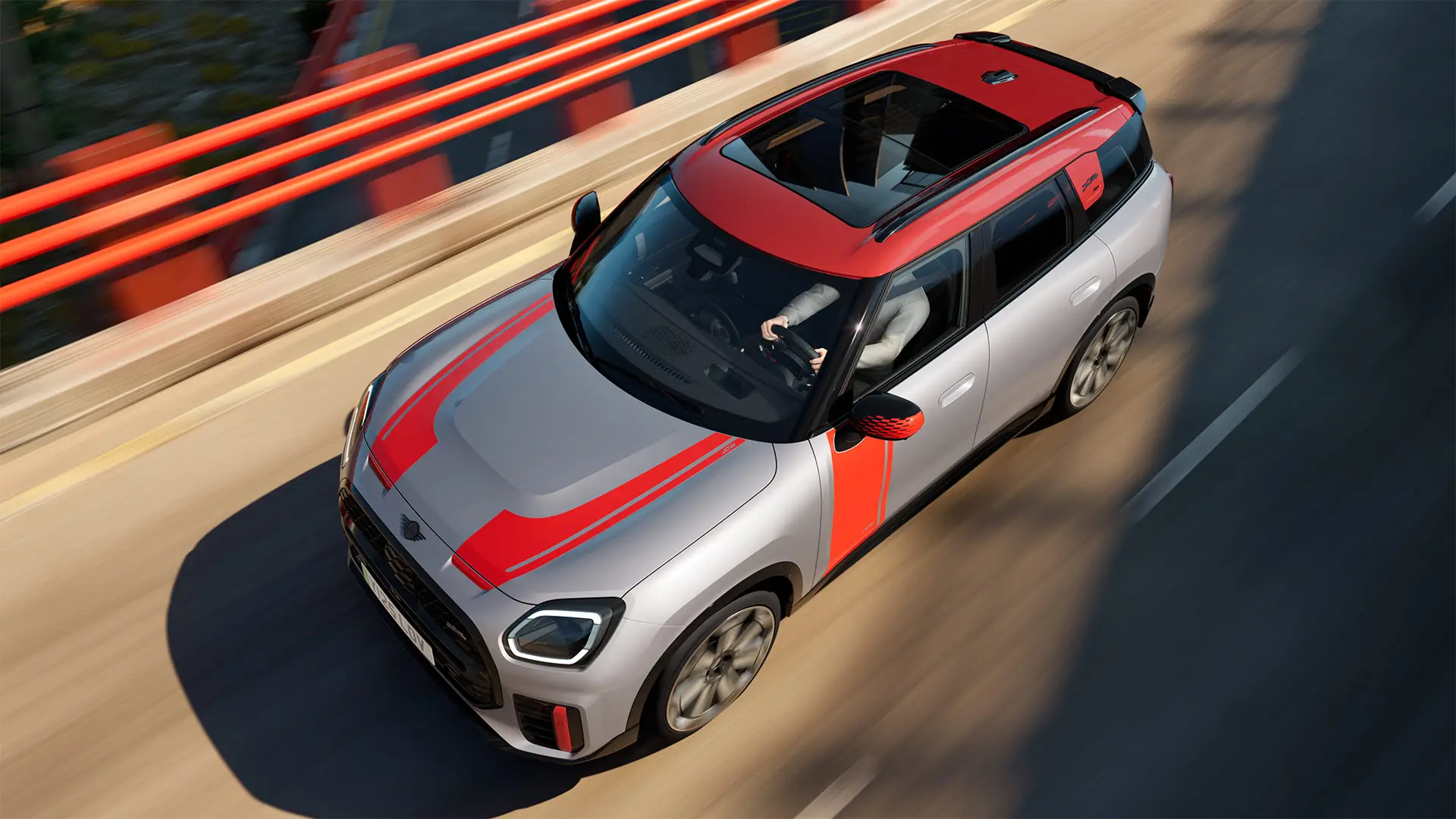 MINI Countryman John Cooper Works in Melting Silver con gusci retrovisori esterni, decalcomanie laterali e sul cofano anteriore, tetto e alette in Chili Red, in marcia lungo una strada cittadina – vista frontale laterale.
