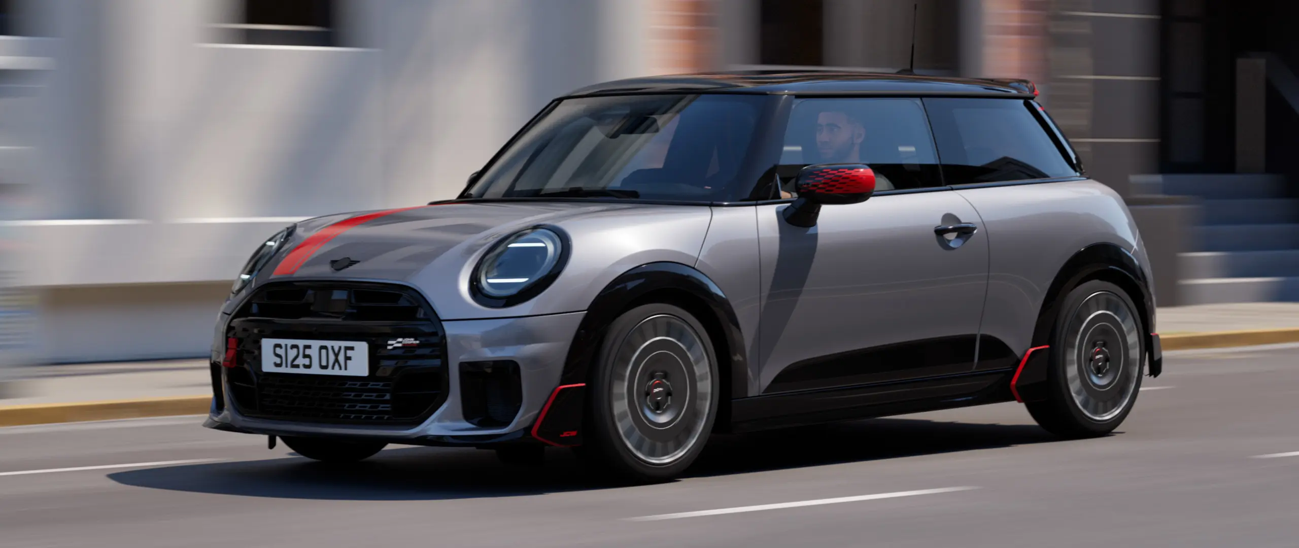 MINI Cooper a 3 porte in Melting Silver con gusci retrovisori esterni John Cooper Works e decalcomania sul cofano anteriore in Chili Red, spoiler su tetto e alettone in Black pastello, mentre percorre una strada cittadina – vista frontale laterale.